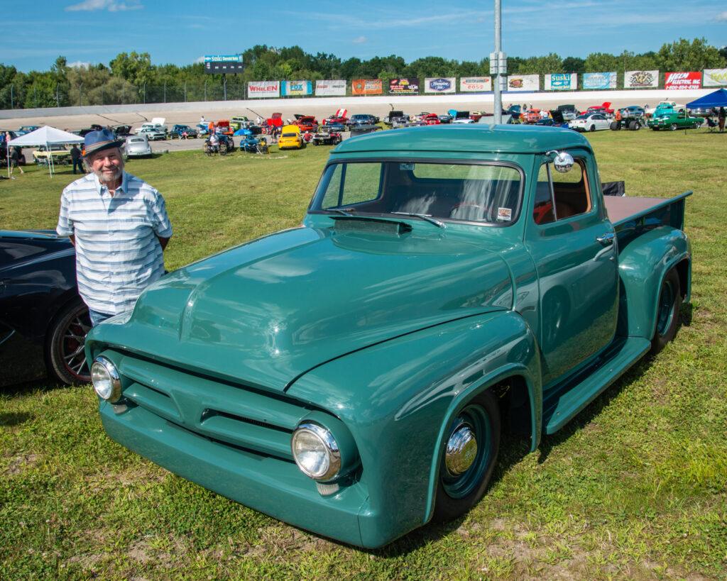 A man stands beside a vintage green pickup truck at a car show.