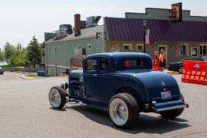 Classic dark blue hot rod car parked on a sunny street near buildings.