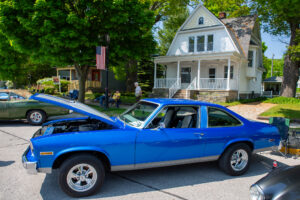 A classic blue muscle car parked in front of a white house on a sunny day.