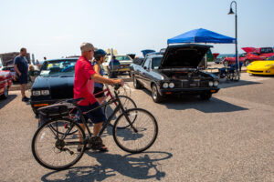 Man in red shirt riding bicycle past classic cars at outdoor event.