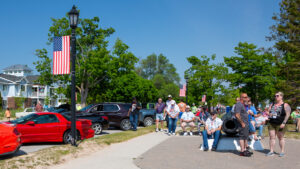 A group of people gathered on a sidewalk near parked cars on a sunny day.