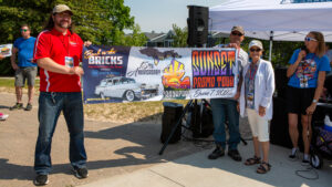 Two men posing by a banner for the Sunset Party 2023 car show with classic cars in the background.