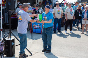 Two men shaking hands during an outdoor event with others watching.