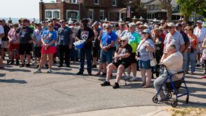 A crowd of people gathered outdoors on a sunny day near brick buildings.