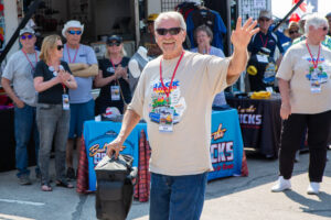 Man waving at an outdoor event, wearing sunglasses and a lanyard.