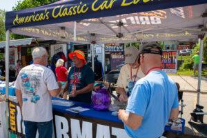 People gather at a classic car event information booth under a tent.