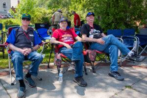 Three older men sitting outdoors on folding chairs, enjoying a sunny day.