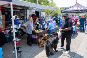 People and dogs gather at an outdoor event with vendor booths under sunny skies.
