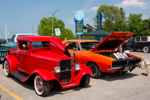 Two classic cars, one red and one orange, displayed outdoors with open hoods.