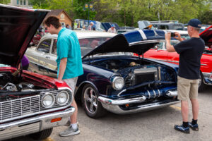 People admiring classic cars at an outdoor car show on a sunny day.