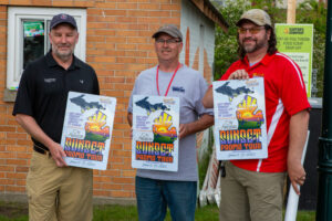 Three men holding event posters, smiling outdoors by a brick building.