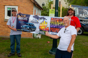Three men proudly display a colorful banner for a car show event outdoors.