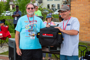 Two men with event badges holding a camera bag outdoors.