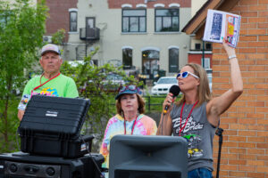 A woman speaks into a microphone at an outdoor event with two others behind her.