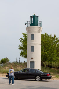 A white lighthouse towering beside a black car and a person sitting nearby.