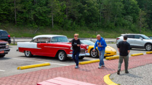 Two men walk near classic and sports cars in a wooded area.