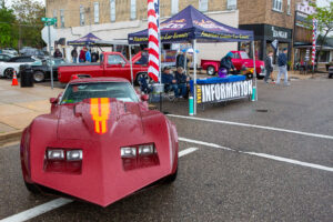 Custom hot rod car at a street car show with an information booth in the background.