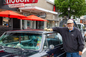 Man leaning on a classic car outside a restaurant with red umbrellas.