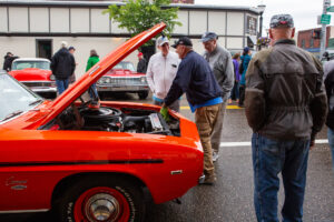 People admire a classic red muscle car with its hood open at an outdoor car show.