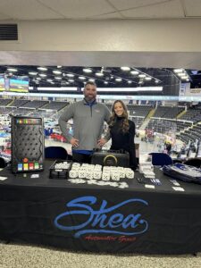 Two people stand behind a Shea-branded table at an indoor event.