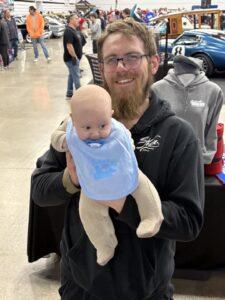 Man holding a baby doll with a big smile at an indoor event.