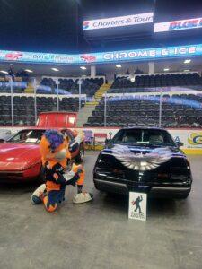 A person in a robot costume poses with two cars in an indoor arena.