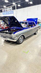 Classic silver car with its hood open at an indoor car show.