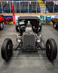 Classic hot rod with exposed engine and vintage design at an indoor car show.