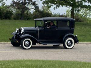 A classic black vintage car driving on a road beside a green field.
