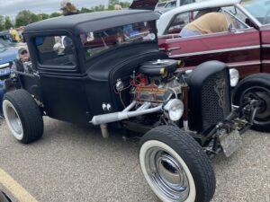 Classic black hot rod with exposed engine and white wall tires at a car show.