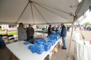 People gather under a tent with tables filled with blue bags for distribution.