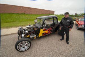 Man in leather jacket stands beside a vintage hot rod car with flame decals.