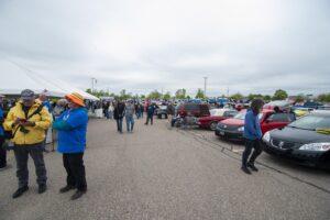 People gather outdoors among parked cars on a cloudy day.