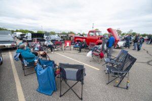 People gather outdoors with vintage trucks and camping chairs in a parking lot event.