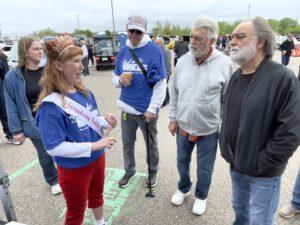 A woman wearing a sash talks to three men outdoors near parked cars.