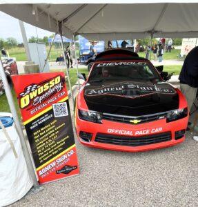 A red sports car displayed under a tent at a car show with a promotional banner beside it.