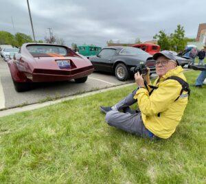 Person in yellow jacket photographing classic cars in an outdoor parking lot.