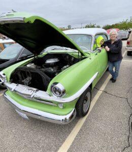 A green vintage car with its hood open at a car show, with a woman standing nearby.