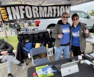 Two people stand behind an information booth at an outdoor event.