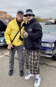 Two men posing together at a car show, smiling and giving thumbs up.