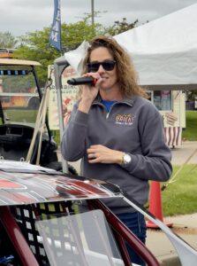 Woman speaking into a microphone at an outdoor event near a car.