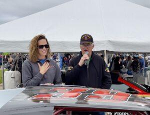 Two people speaking at an outdoor event under a white tent.