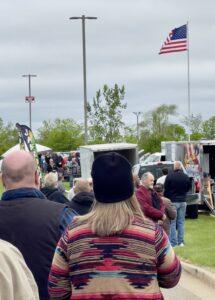 A crowd gathers outdoors under cloudy skies with flags and trailers visible.