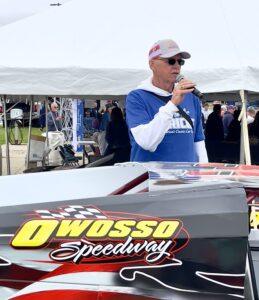 Man speaking at Owosso Speedway event with race car nearby.