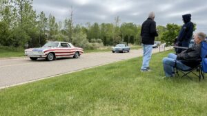 Two vintage cars driving on a road with a person standing on the grass beside them.