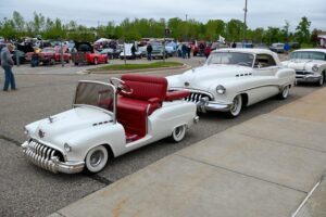 A miniature white car towing a classic white Buick on a road.