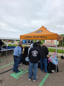 People gather under an orange Coffee Beanery tent at an outdoor event.