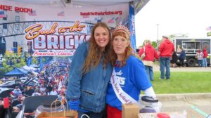 Two women at a community event, one wearing a sash and crown.
