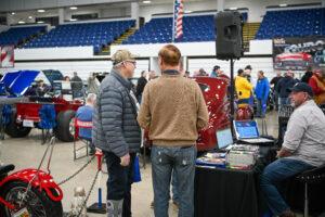 Two men conversing at a vendor table inside a large indoor venue.