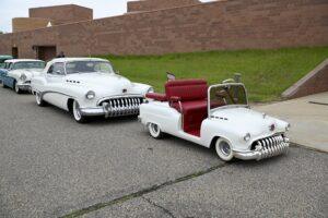 A white vintage car and a smaller matching toy car on a paved path.